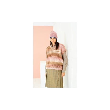 A woman wearing a pink and brown striped knit sweater vest over a grey long-sleeve top, paired with a brown skirt and a pink knit beanie, standing against a minimalist pink and white background.