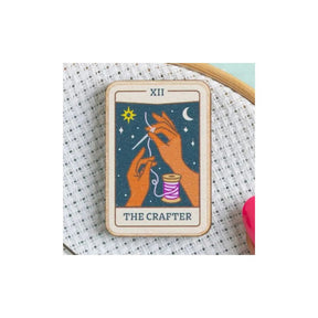 A tarot card enamel pin depicting 'The Crafter' (XII) with illustrated hands, a spool of thread, and a star against a dark blue background, shown on a white cross-stitch canvas.