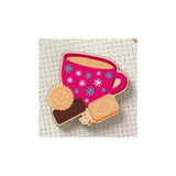 A small embroidered patch or applique featuring a pink cup with blue and white starburst patterns, alongside two cookies, shown on a white textured fabric background.