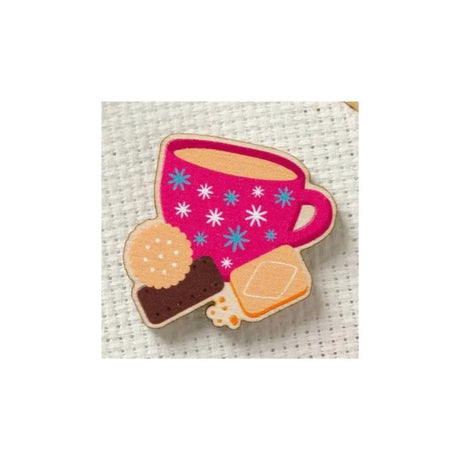 A small embroidered patch or applique featuring a pink cup with blue and white starburst patterns, alongside two cookies, shown on a white textured fabric background.