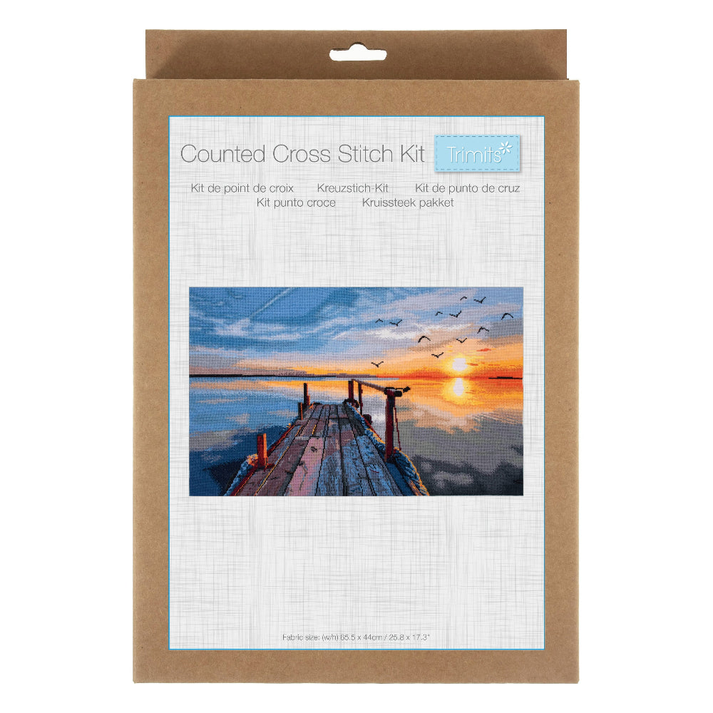 A counted cross stitch kit in a brown kraft paper sleeve with a white front label showing a sunset dock over water and the text 'Counted Cross Stitch Kit' and 'Trolli'.