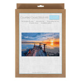 A counted cross stitch kit in a brown kraft paper sleeve with a white front label showing a sunset dock over water and the text 'Counted Cross Stitch Kit' and 'Trolli'.