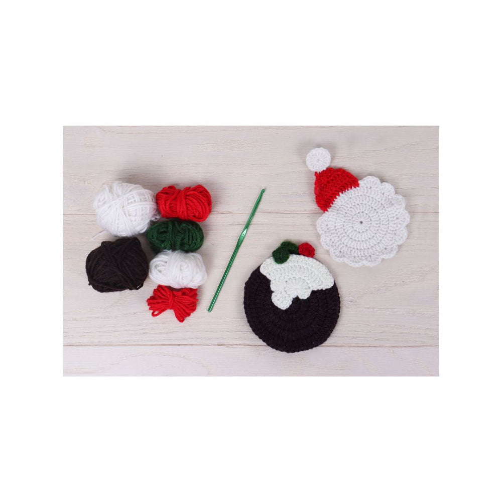 A flat-lay of crocheted Christmas-themed items on a light wood background, including a white crocheted Santa hat, a white and red Santa face, a black and white pudding, several balls of yarn in white, red, green, and black, and a green crochet hook.