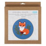 An embroidery hoop kit in its original brown kraft paper packaging, featuring a printed design of an orange and white fox on a blue background with green leaves.