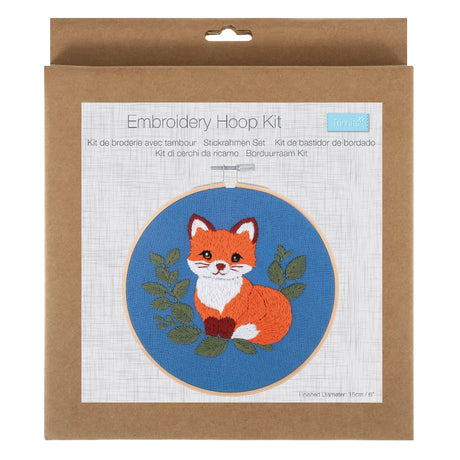 An embroidery hoop kit in its original brown kraft paper packaging, featuring a printed design of an orange and white fox on a blue background with green leaves.