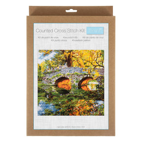 A counted cross stitch kit in its original brown kraft paper packaging, featuring a colorful illustration of a stone bridge over a reflective pond surrounded by autumn trees.