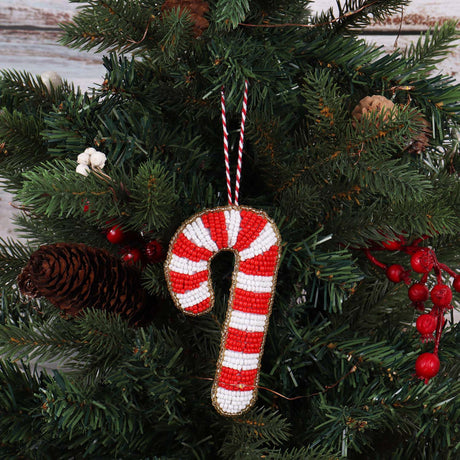 A handmade beaded Christmas candy cane ornament hanging on a green pine tree with red berries and a pinecone.