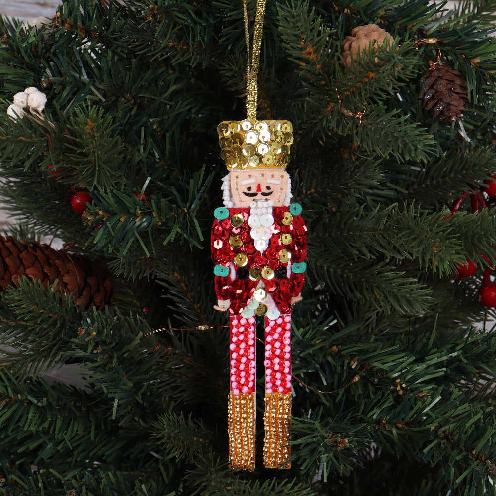 A sequined nutcracker Christmas ornament hanging from a gold ribbon against a green Christmas tree background.