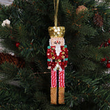 A sequined nutcracker Christmas ornament hanging from a gold ribbon against a green Christmas tree background.