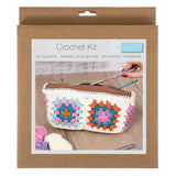 A crocheted zipper pouch shown inside its retail packaging box. The box is kraft brown with a printed front photo of the finished multicolor pouch, crochet hooks, and yarn balls.