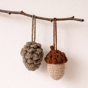Two crocheted acorn and pinecone hanging ornaments suspended from a natural twig against a plain off-white wall.