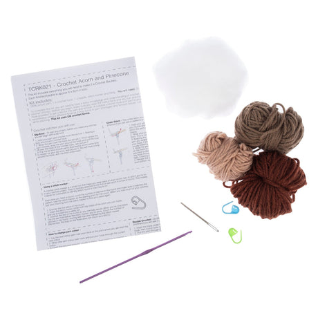 Crochet acorn and pinecone kit components on a white background, including a printed instruction sheet, white fiber ball, four yarn balls in beige, taupe, and brown, a metal hook, a blue stitch marker, a green stitch marker, and a purple crochet hook.