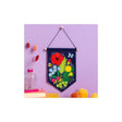 A colourful felt wall hanging banner with embroidered flowers and leaves, suspended from a wooden dowel on a lavender wall. The banner features a navy blue border and a vibrant floral design with red, yellow, green, and blue details.