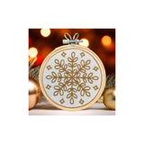 A circular embroidery hoop containing a gold snowflake and geometric pattern on white fabric, shown against a warm blurred holiday background with lights and gold ornaments.