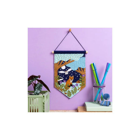 A decorative fabric wall hanging banner with a woodland scene featuring a fox, moon, stars, and trees, suspended by a dark cord from a wooden rod against a lavender wall. The banner is styled in a room setting with a small plant and desk accessories.