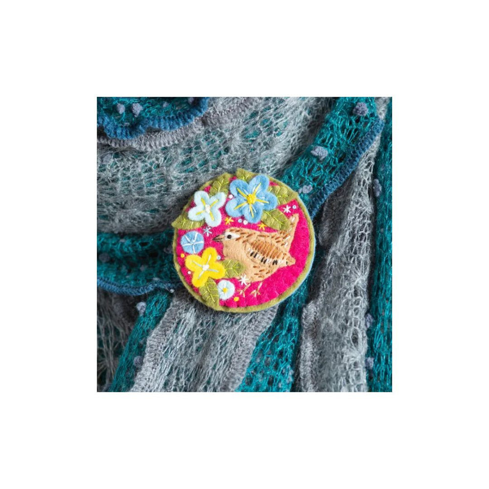 A round embroidered patch featuring a brown bird and colourful flowers on a pink background, pinned onto a grey and teal knitted sweater.