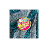 A round embroidered patch featuring a brown bird and colourful flowers on a pink background, pinned onto a grey and teal knitted sweater.
