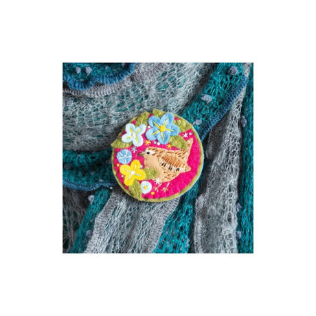 A round embroidered patch featuring a brown bird and colourful flowers on a pink background, pinned onto a grey and teal knitted sweater.