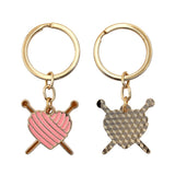 Two gold keychains with heart and knitting needle charms on a white background
