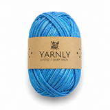 A ball of Yarnly Lustre T-shirt yarn with a brown paper label, featuring a shimmering blend of blue threads, on a white background.